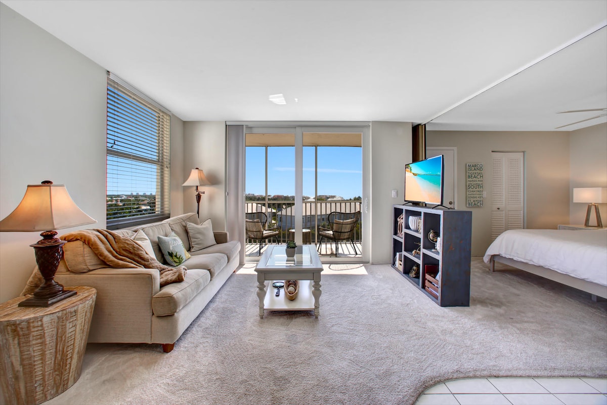 A bright living area features a comfortable sofa flanked by table lamps. A flat-screen TV is positioned on a shelving unit. Floor-to-ceiling sliding glass doors lead to a balcony with outdoor seating, providing views of the surrounding area and natural light.