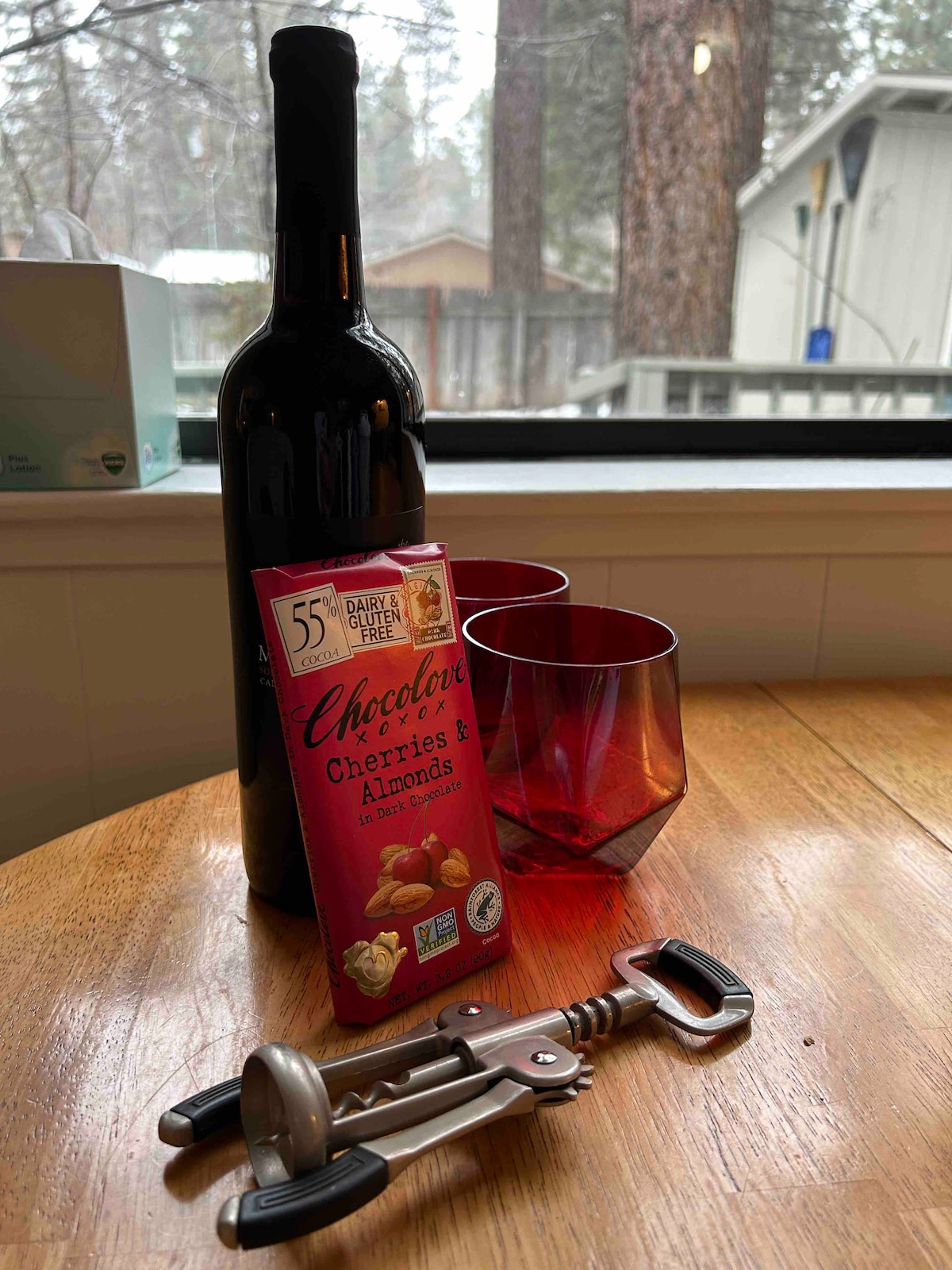 A bottle of red wine is placed on a wooden table alongside two red glasses. A box of chocolate-covered cherries and almonds is visible, along with a corkscrew and bottle opener. The background features large windows with a view of trees, creating a cozy indoor setting.