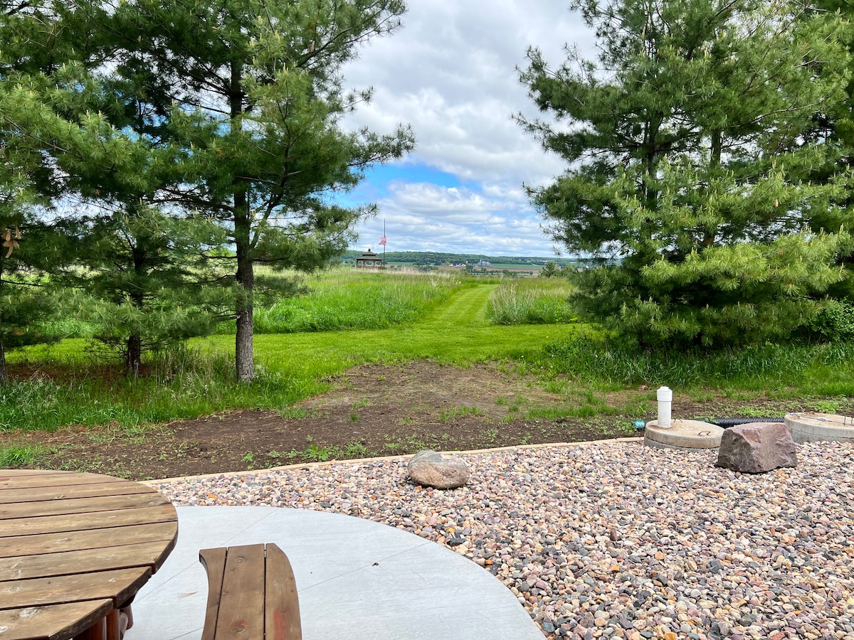 A peaceful outdoor scene features a well-maintained grassy area framed by tall pine trees. A fire pit with seating is visible in the foreground, while the landscape opens up to distant hills under a partly cloudy sky.