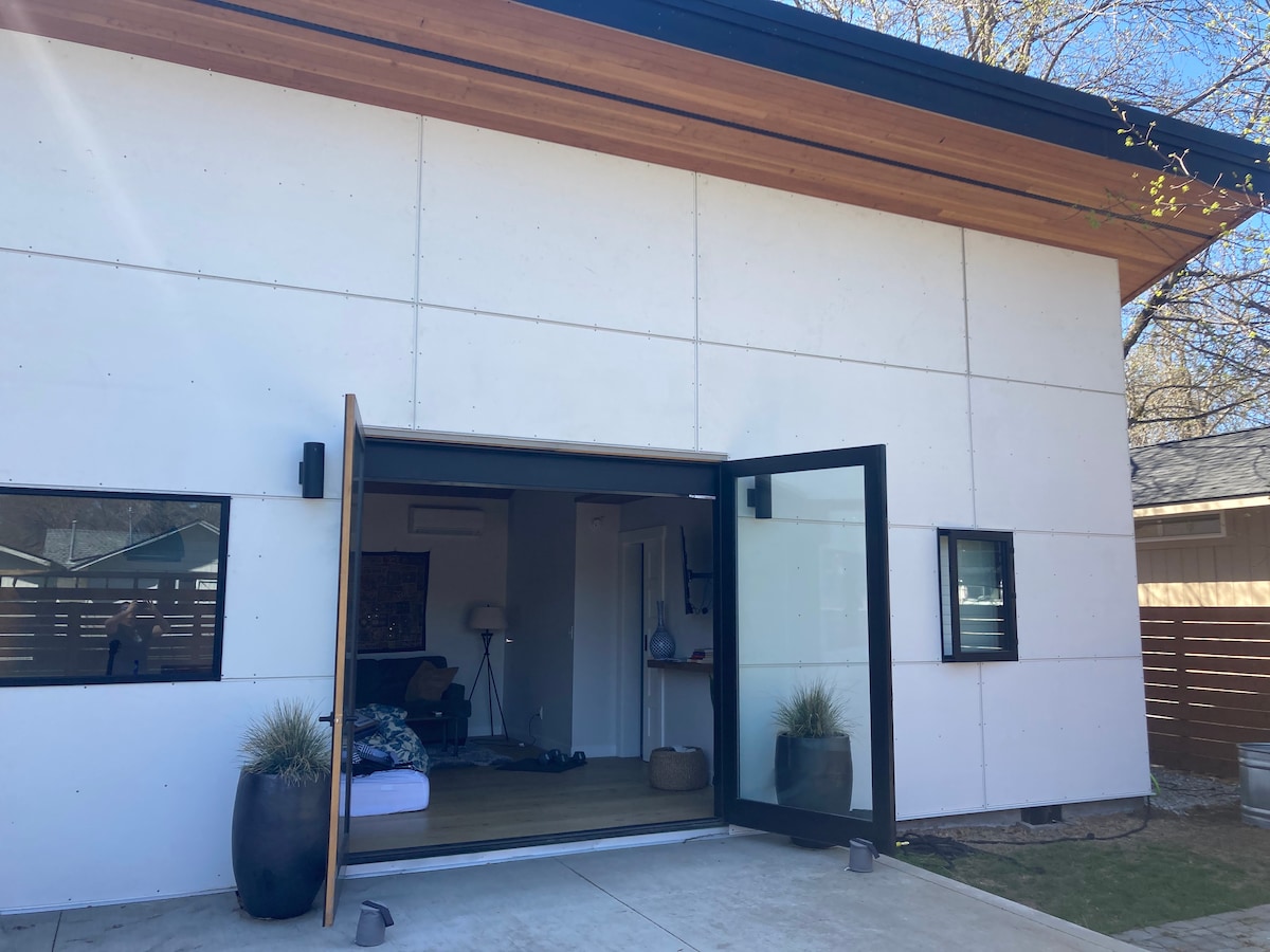 The exterior of the bungalow features a contemporary design with large, open glass doors that invite natural light. Two windows are visible beside the entrance, and minimal landscaping, including potted plants, adds to the serene ambiance.