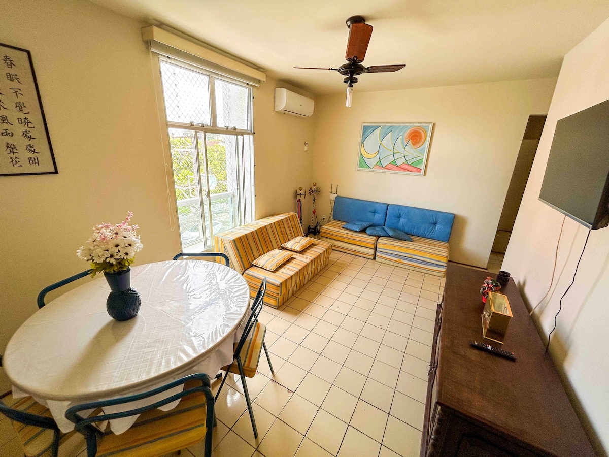 A bright living area features two striped sofa beds and a round dining table set with chairs. A colorful wall art piece is displayed, and light enters through a large window. A ceiling fan and air conditioning are present for comfort.