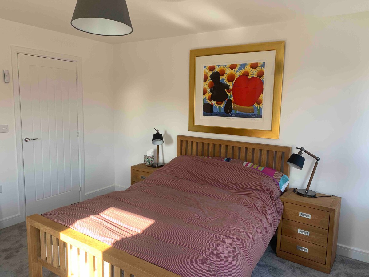 A cozy bedroom is furnished with a wooden bed featuring a striped duvet. Two bedside tables with lamps sit on either side. A colorful framed artwork adorns the wall above the bed. Natural light filters in through a nearby window, highlighting the soft carpet.