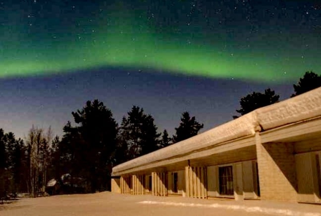 A tranquil scene captures the northern lights illuminating the night sky in shades of green above a low building. Snow blankets the ground, with silhouettes of trees on the horizon creating a serene backdrop.