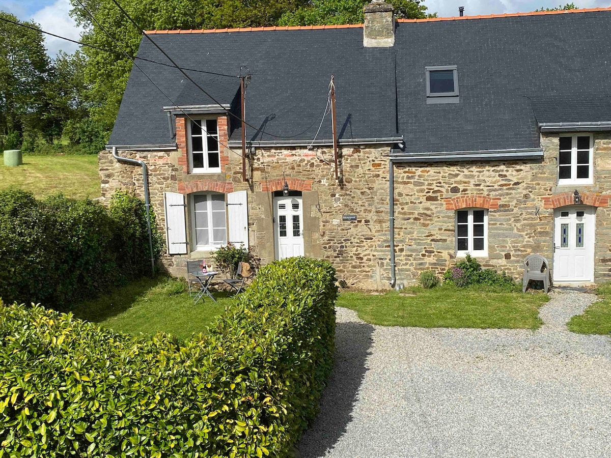 The exterior of Le Cottage is characterized by natural stone walls and a slate roof. A small outdoor seating area with a bistro table and chairs is set in front of the cottage, surrounded by neatly trimmed hedges. Lush greenery is visible in the background.
