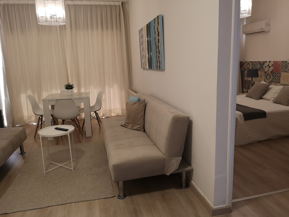 An open plan living area features a light-colored sofa and a small round table surrounded by four chairs. A textured rug lies beneath, and a painting adds a touch of color to the wall. A separate bedroom is visible through a sliding door.