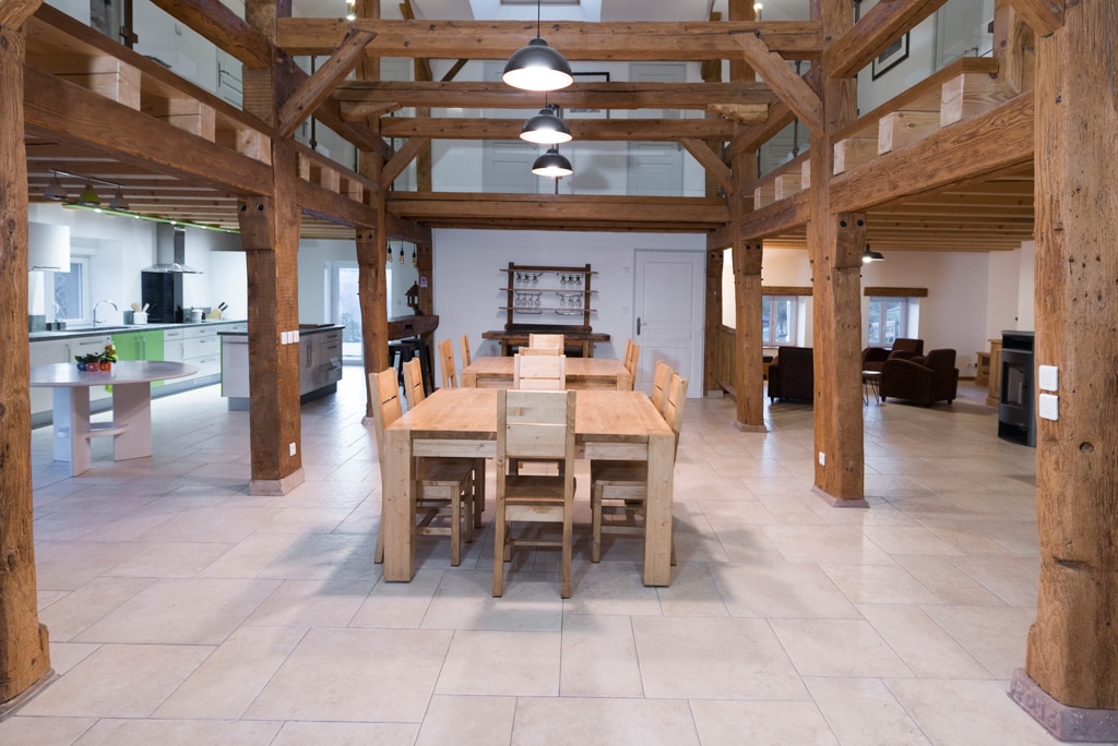 A spacious dining area features a large wooden table surrounded by simple wooden chairs. Exposed wooden beams support the high ceiling, adding character to the room. The adjacent kitchen area is visible, equipped with modern appliances and an inviting layout.
