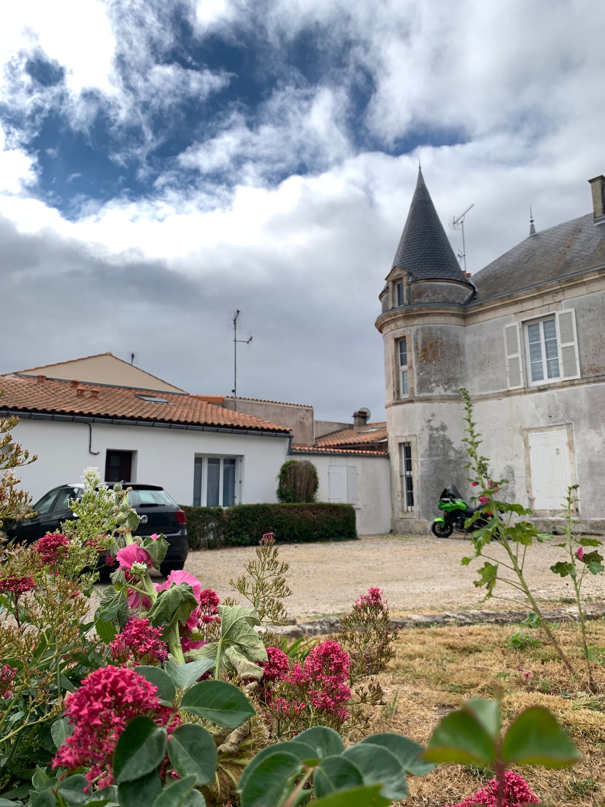 A charming house is highlighted alongside a historic structure featuring a prominent tower. The gravel courtyard is dotted with green shrubs and blooming flowers, with a parked vehicle visible. Overcast skies create a soft ambiance above the property.