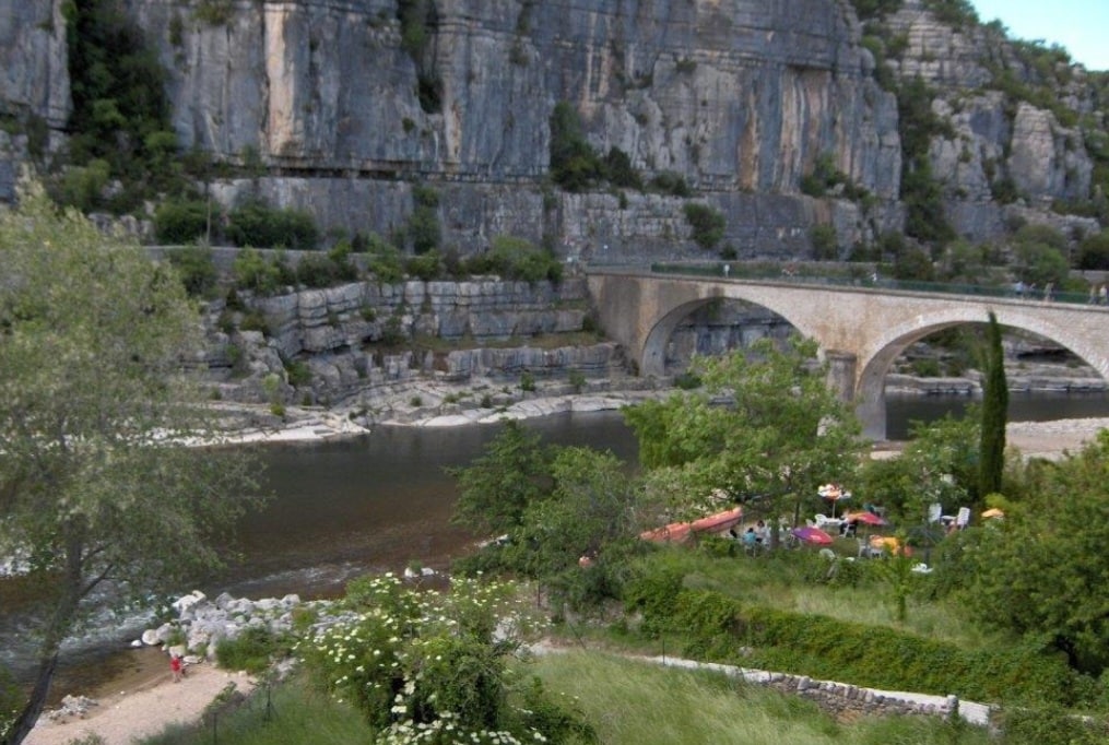 A scenic view of the river Ardèche showcases its tranquil waters beside rocky cliffs. A stone bridge spans the river, while colorful umbrellas and seating areas appear along the shoreline, surrounded by lush greenery. The peaceful landscape emphasizes the natural beauty of the area.