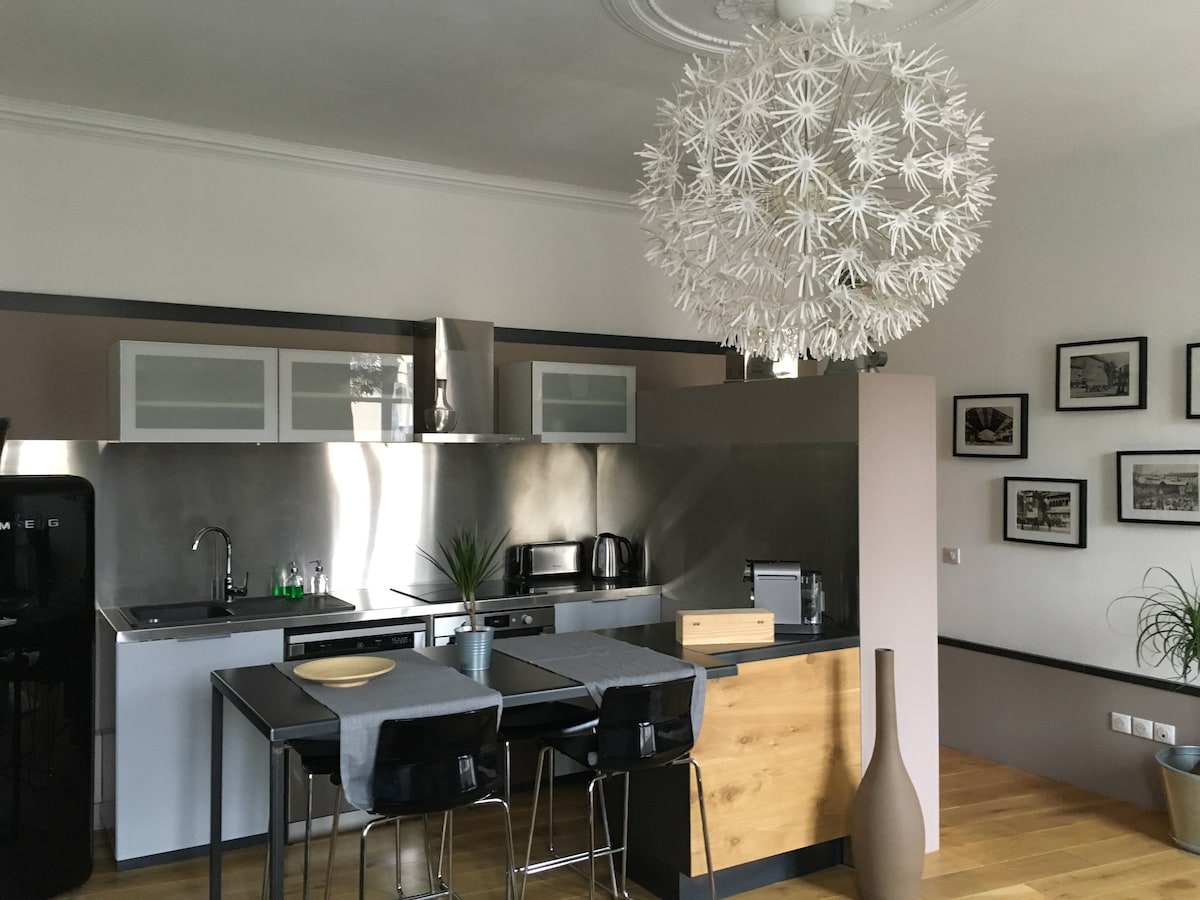 A modern kitchen area features sleek metal cabinetry and an island with two stools. A striking floral pendant light hangs overhead, casting unique shadows. Wall art and potted plants add to the contemporary design, enhancing the overall ambiance.