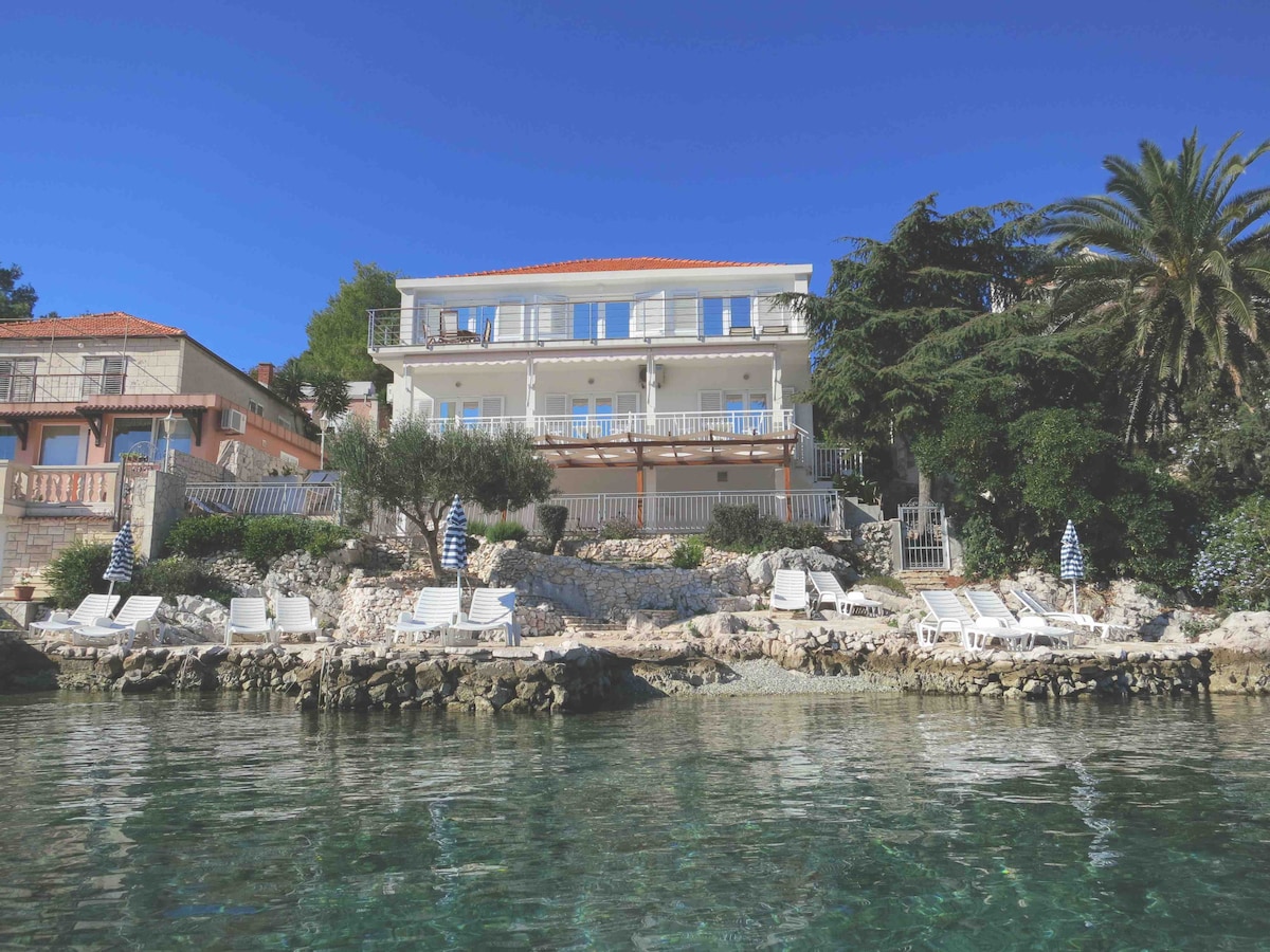 A three-story building is situated by the sea, featuring balconies that overlook the water. Sun loungers are arranged along the rocky coastline, providing space for relaxation. Lush palm trees and olive trees are visible, adding to the natural surroundings of the property.