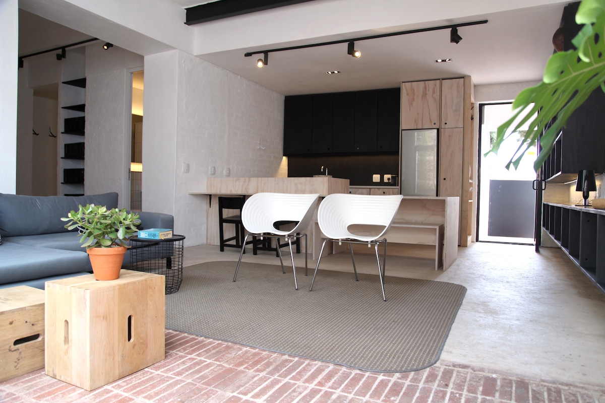 An open-plan living area is visible, featuring a modern kitchen with sleek cabinetry and a stylish lounge space. Two white chairs are positioned around a coffee table, and a potted plant adds a touch of greenery. Natural light illuminates the interior through the front door.