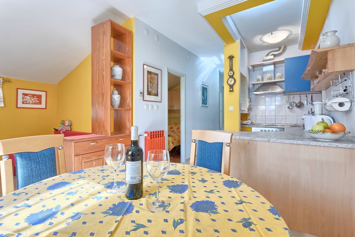 A dining area features a round table set with blue and yellow patterned tablecloth, accompanied by four wooden chairs. A bottle of wine and glasses are placed in the center. The kitchen is seen in the background, showcasing blue cabinetry and cooking appliances.