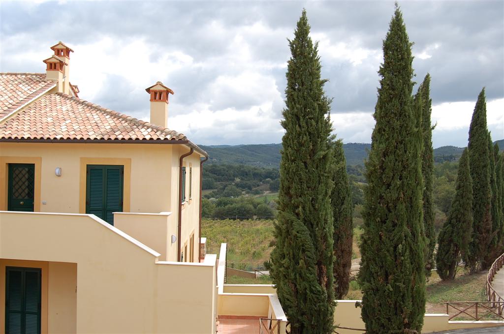 The exterior of the duplex maisonette features a warm yellow façade and a tiled roof, surrounded by tall cypress trees. The hills of Tuscany are visible in the background, creating a serene landscape under a cloudy sky.