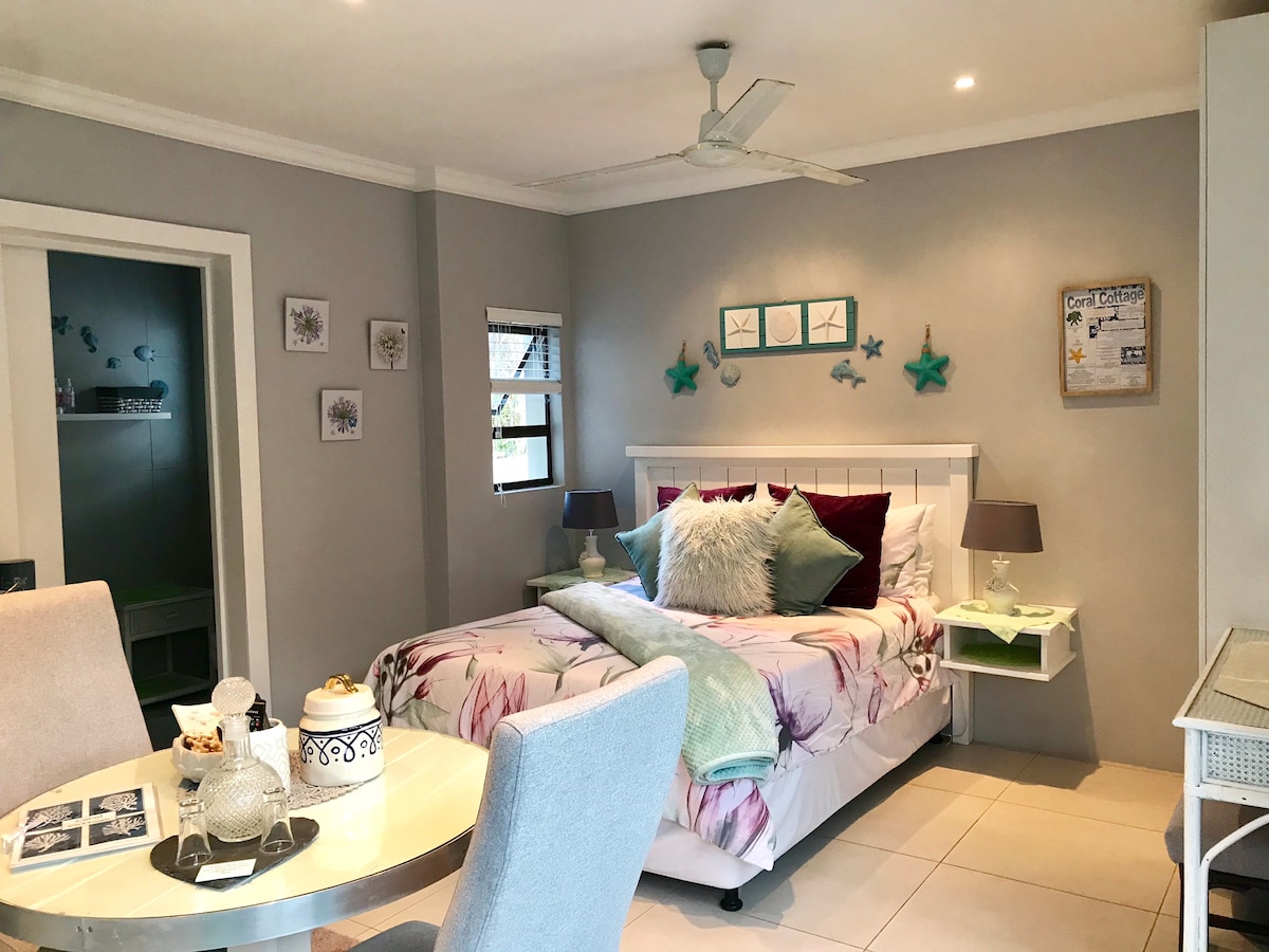 A cozy bedroom is presented with a spacious bed adorned in floral bedding, accompanied by soft pillows. A small dining table with two chairs is visible, complemented by a fan and decorative wall art. Natural light filters through a nearby window, enhancing the calming ambiance.