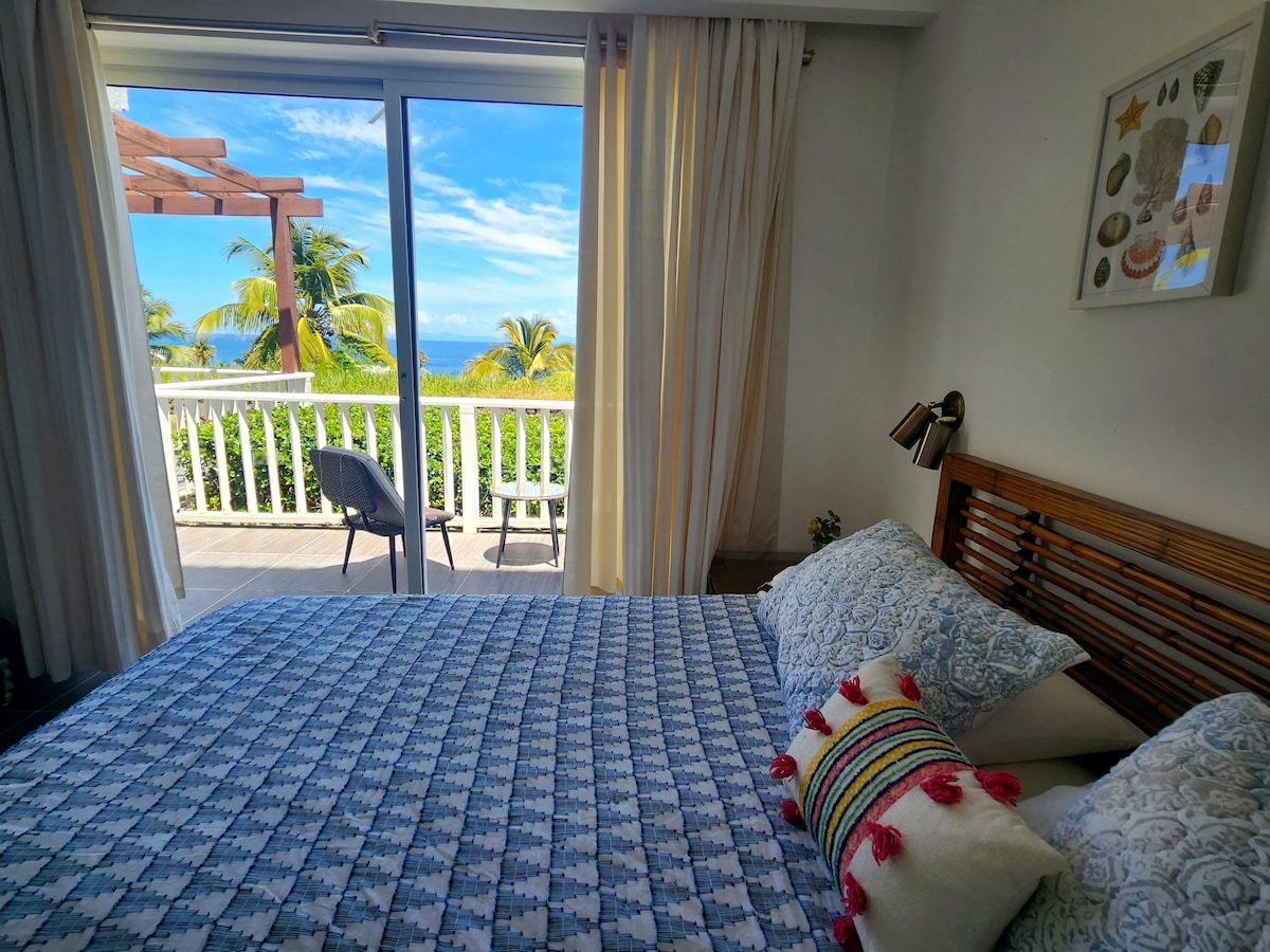 A well-lit bedroom features a spacious bed adorned with a patterned blanket and vibrant decorative pillows. Light flows in through sliding glass doors, which lead to a private balcony overlooking lush greenery and the ocean in the distance. A modern lamp stands beside the bed.