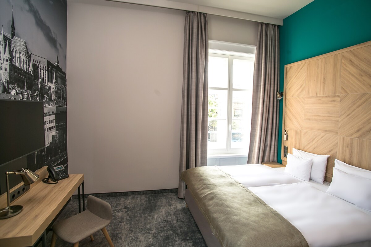 A well-lit hotel room features a spacious bed with crisp white linens and a light-colored blanket. A large window allows natural light to enter, flanked by gray curtains. A desk with a telephone is positioned nearby, while a black-and-white cityscape is displayed on one wall.