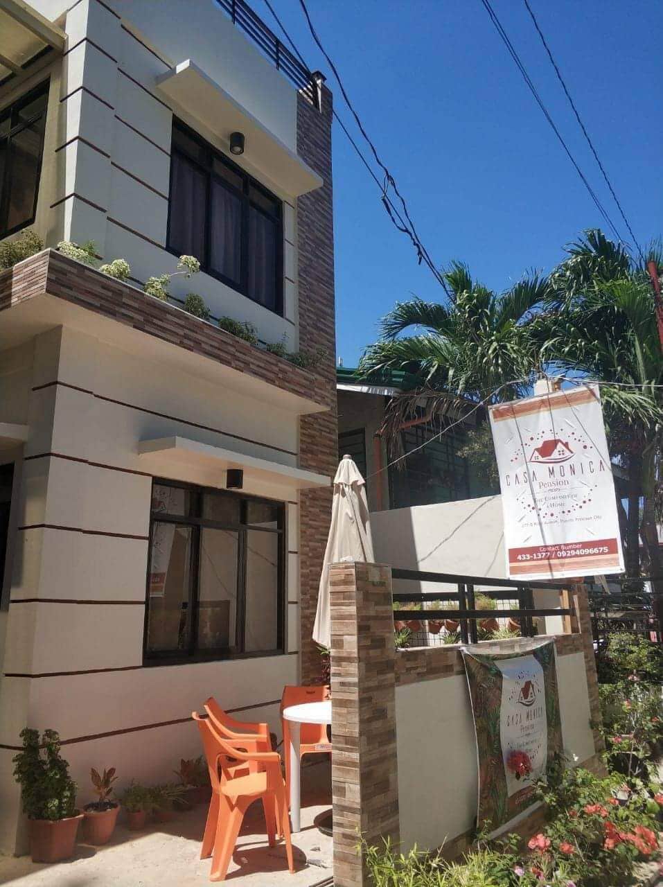 The exterior of the pension house is presented, featuring a modern architectural design with light-colored walls and a decorative stone accent. A small patio area is visible, displaying orange chairs and a table, with potted plants and a sun umbrella enhancing the outdoor space.
