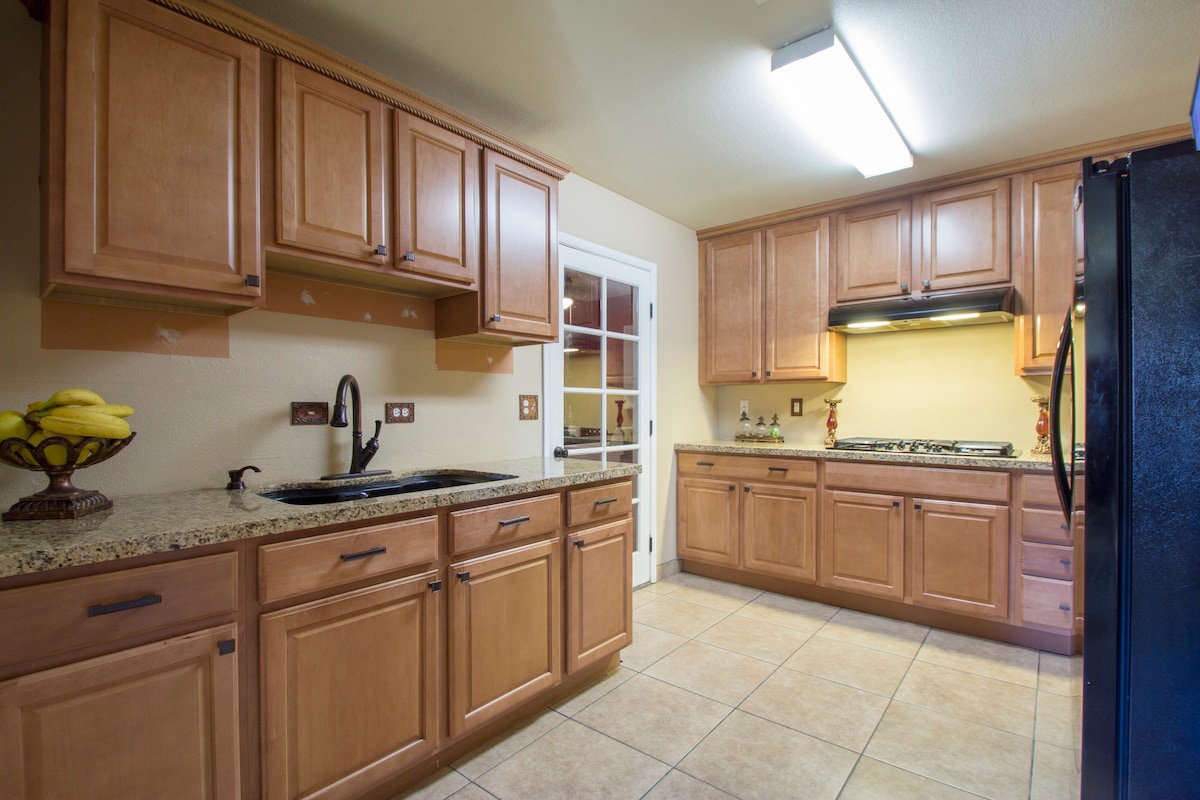 A modern kitchen is featured with wooden cabinetry and granite countertops. A black refrigerator is present alongside a sink and a gas range. Natural light is allowed in through a nearby door, leading to a well-organized and functional space.