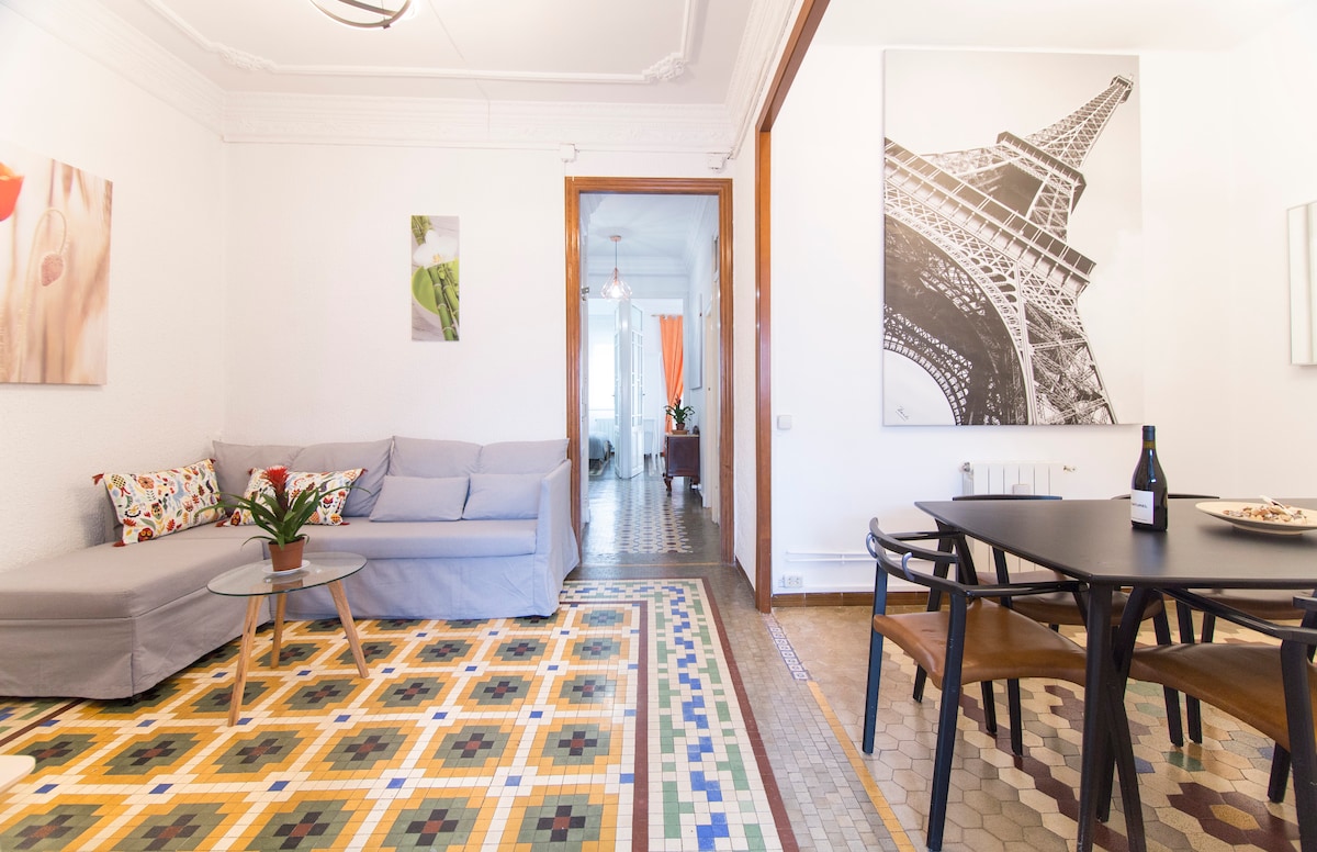 A spacious living area features a light grey sofa facing an elegant black dining table with four chairs. Decorative wall art includes floral prints and a black-and-white photograph of the Eiffel Tower. The room boasts intricate hydraulic tile flooring in various colors.