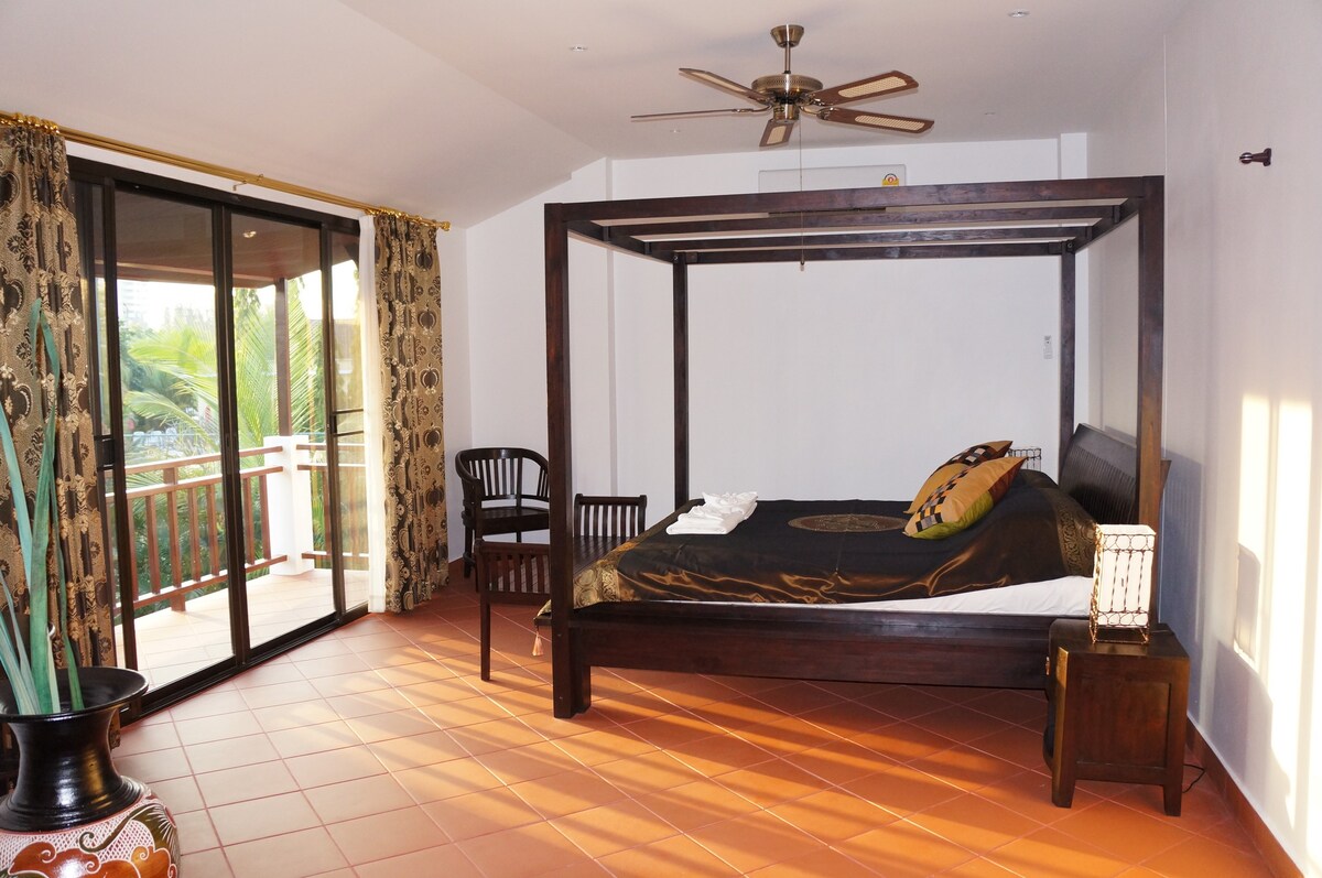 A spacious bedroom features a four-poster bed with dark bedding. Large glass doors open to a balcony, allowing natural light to fill the room. A wooden chair, a decorative table, and a potted plant are visible, enhancing the open and airy feel.