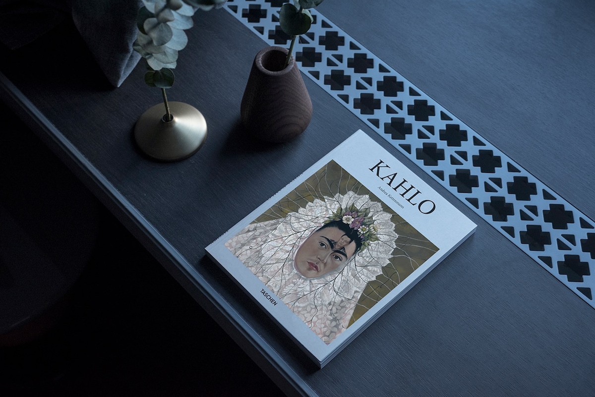 A stylish tabletop displays a book titled 'KAHLO,' featuring elegant artwork and floral accents. A small vase and a decorative candle holder, both in muted tones, add to the modern decor. A geometric pattern runs along the edge of the table, enhancing the visual appeal.