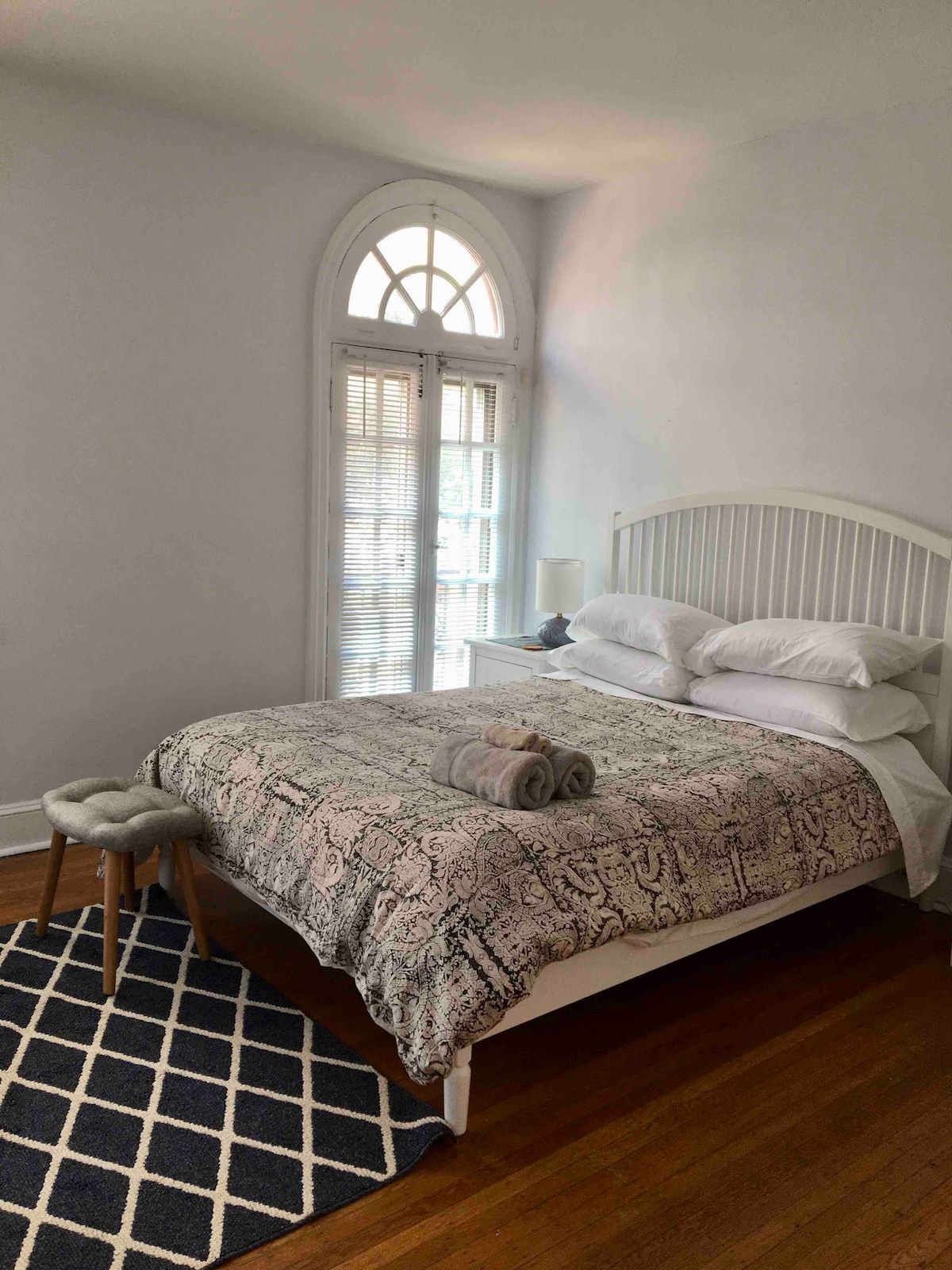 A bright bedroom features a spacious bed adorned with decorative bedding. A rolled towel is placed on top, and a small stool sits nearby. Natural light filters through a large arched window with shutters, illuminating the hardwood floor.