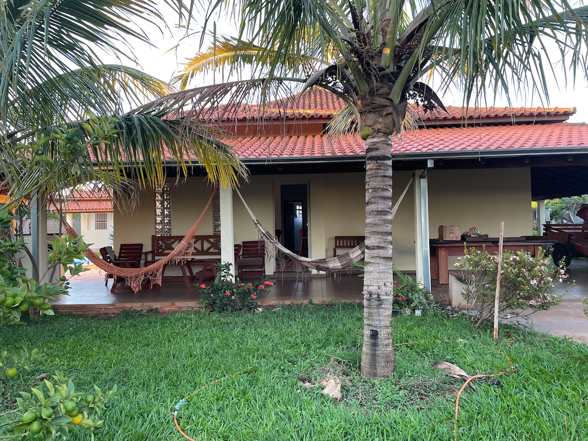 A cozy exterior view of a home surrounded by well-maintained green grass and tropical plants. A porch features comfortable wooden seating, with hammocks hanging beside it. The red-tiled roof complements the warm tones of the house, creating a relaxed atmosphere.