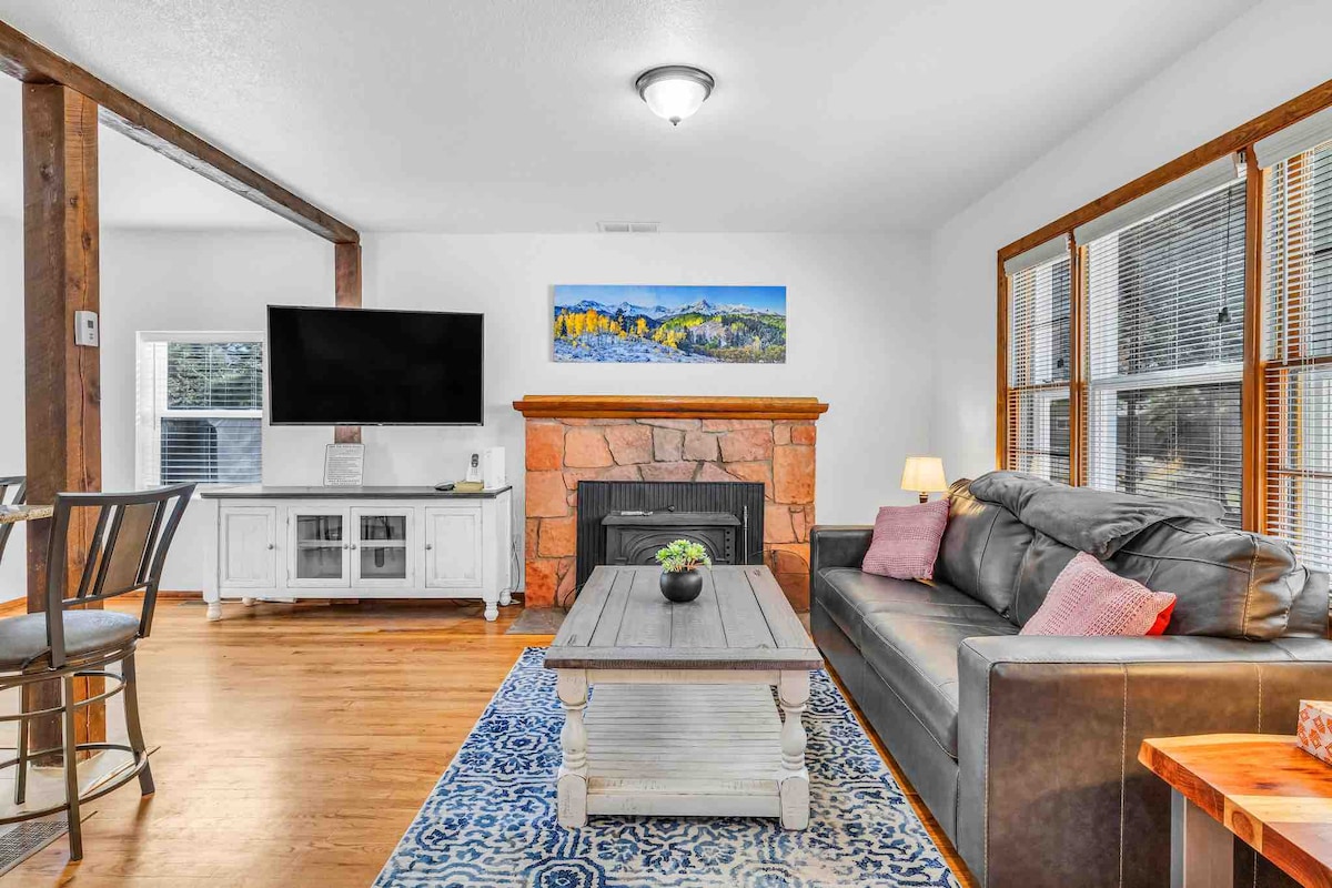 A comfortable living area features a dark leather sofa, accented with pillows, facing a large flat-screen television. A stone fireplace adds warmth to the space, which is illuminated by natural light from the nearby windows. A decorative rug and a wooden coffee table complete the inviting setting.
