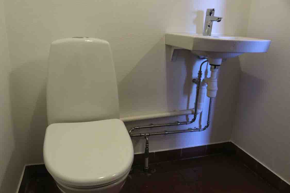 A clean and functional bathroom is depicted, featuring a white toilet positioned beside a compact sink. The sink connects to visible plumbing beneath, and the walls are finished in a light hue. The floor is tiled in a dark color, complementing the overall design.