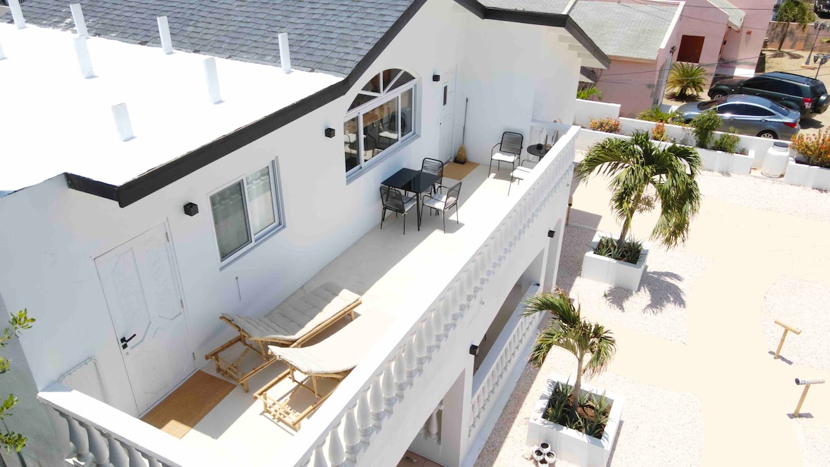 A spacious balcony overlooking the surroundings is furnished with two lounge chairs and a small table. The area is bordered by elegant railings and features decorative plants, creating an inviting outdoor space for relaxation.