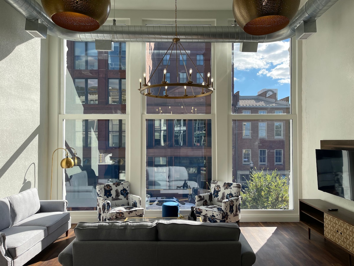 A bright living area is highlighted by large windows allowing natural light to flood the space. Two patterned chairs are positioned near a coffee table, with a gray sofa facing a mounted television. A modern chandelier adds a stylish touch, complementing the urban view.