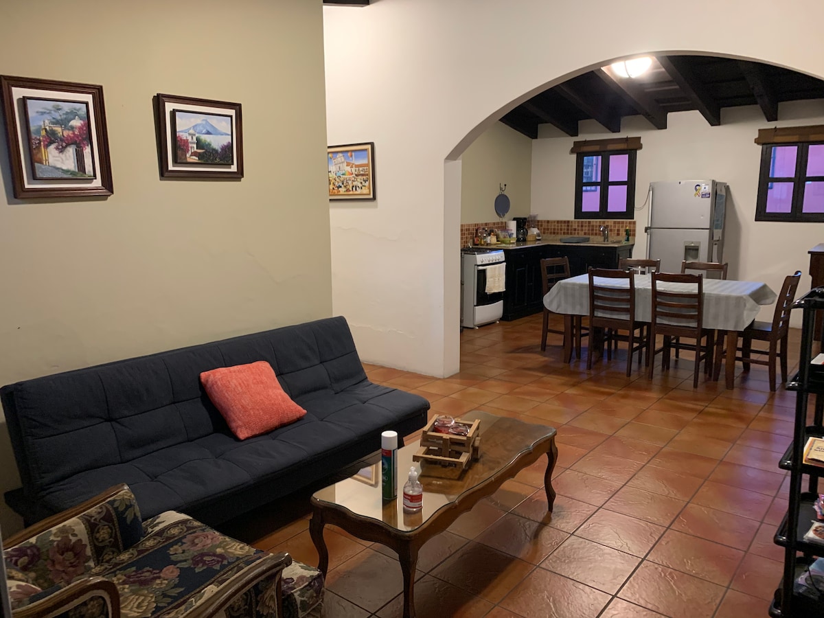 A spacious living area is shown featuring a dark blue sofa with an orange accent pillow, paired with a patterned armchair. An archway leads to a dining area, furnished with a wooden table and chairs. Original artwork adorns the walls, and a kitchen is visible in the background.