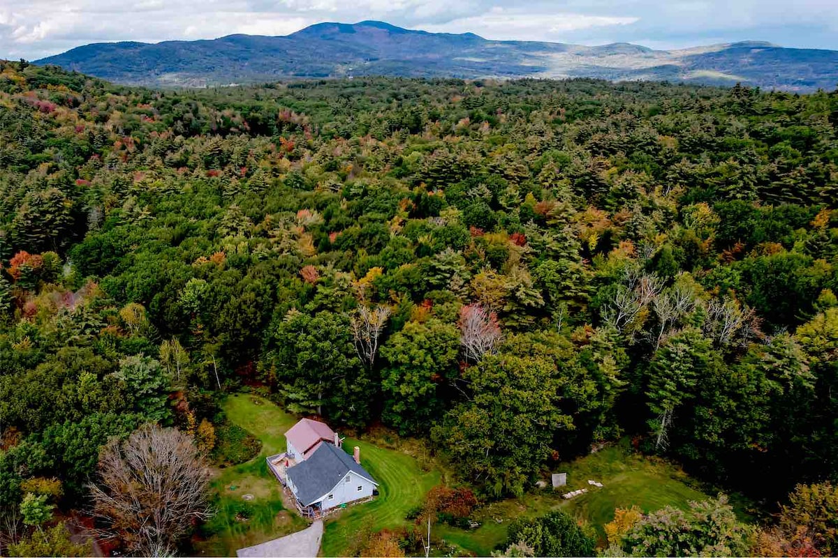 An aerial view captures the home surrounded by lush greenery and diverse tree cover, showcasing a blend of evergreens and deciduous trees. Rolling hills and distant mountains provide a scenic backdrop, while the well-maintained lawn and wooded surroundings offer a sense of seclusion and tranquility.