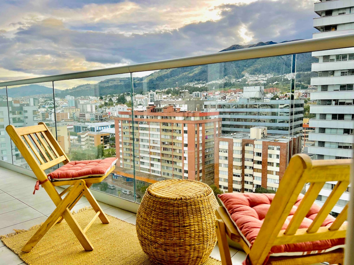 A private balcony features two wooden folding chairs with cushions and a round woven table, offering views of the cityscape and surrounding mountains. The glass railing provides an unobstructed sightline of the urban landscape beneath a cloudy sky.