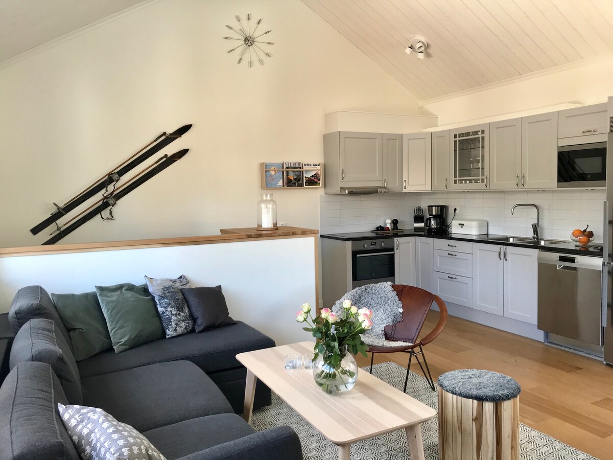 A well-equipped kitchen area features modern appliances and sleek cabinetry, complemented by a cozy living space. A gray sectional sofa is arranged beside a light wooden coffee table. Decorative elements include a vase with fresh flowers and vintage skis mounted on the wall.