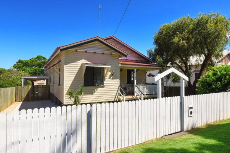 A charming cottage is framed by a white picket fence, showcasing a welcoming front porch with seating. Lush greenery surrounds the home, and clear blue skies enhance the overall appeal. The house features a traditional design with a sloped roof and ample windows.