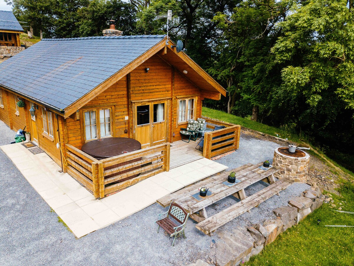 The log cabin, surrounded by green foliage, displays a spacious wooden deck with outdoor seating and a private hot tub. A stone seating area is visible on the right, featuring a rustic table and chairs. The slate roof and wooden paneling give a charming, rustic appearance.