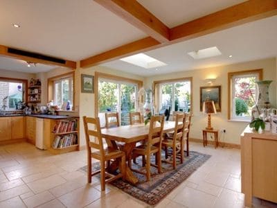 A spacious dining area features a large wooden table surrounded by six chairs. Natural light floods in from multiple windows overlooking the garden. The kitchen is visible in the background, complemented by wooden cabinetry and an inviting atmosphere.