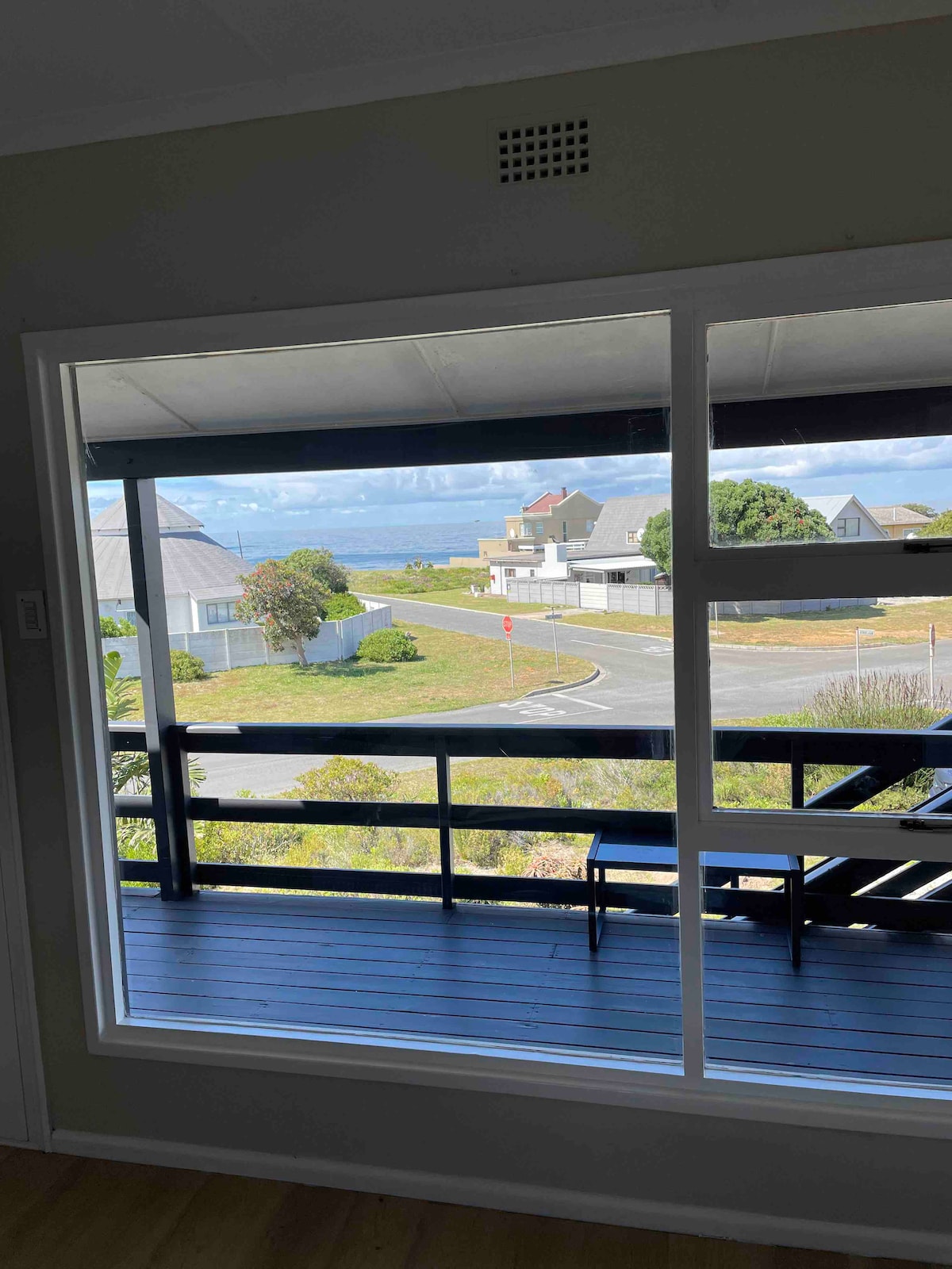 A large window frames a view of the surrounding neighborhood, with a glimpse of the sea beyond. The deck area features wooden flooring, and a chair is positioned near the edge, allowing for relaxation while enjoying the scenery.