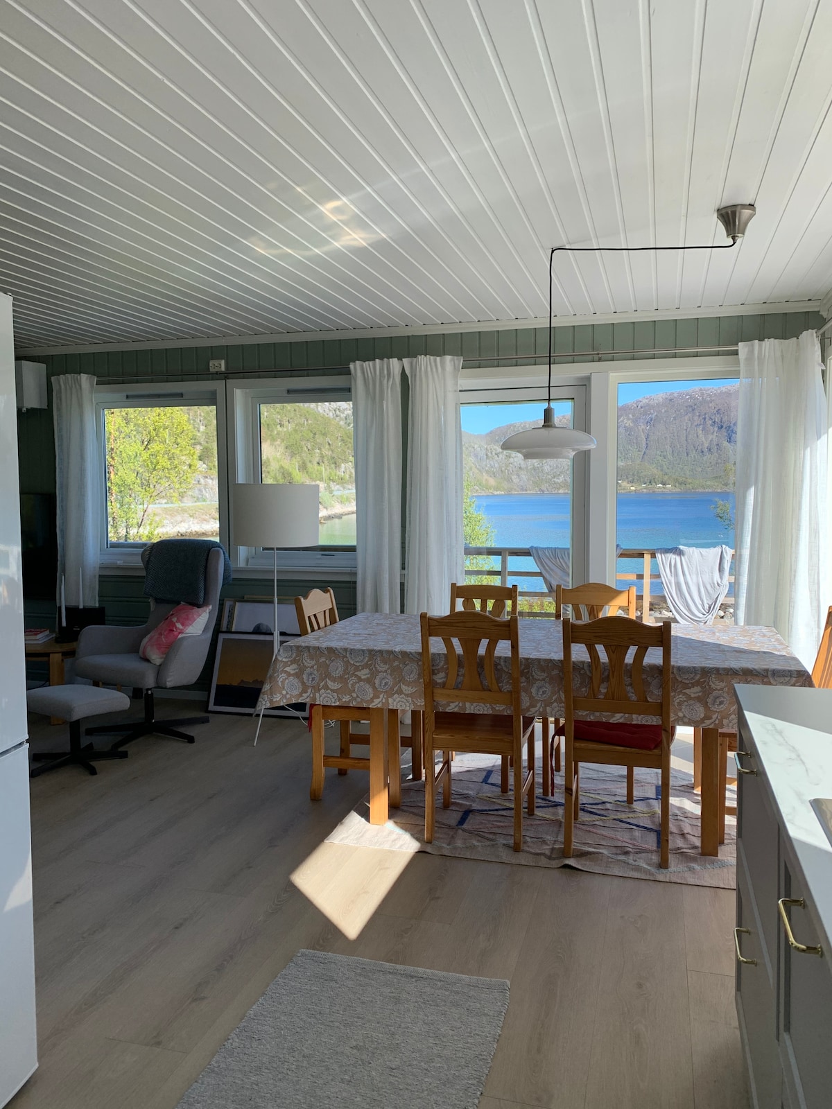 A dining area features a wooden table surrounded by six chairs, illuminated by natural light from large windows. A comfortable armchair is positioned nearby, while scenic views of the lake and mountains are visible through the glass. Light-colored curtains frame the windows.