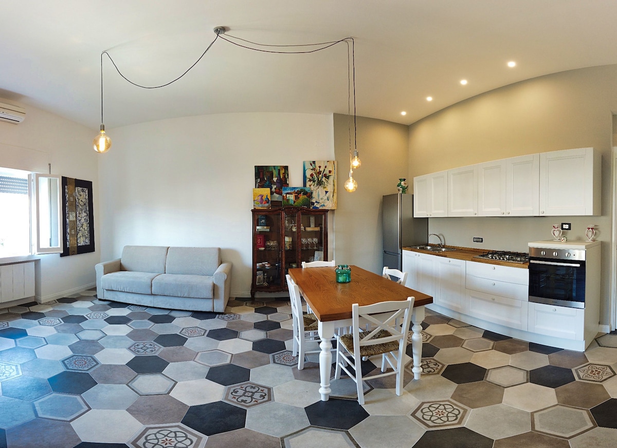 A spacious living area features a light-coloured sofa and a wooden dining table surrounded by white chairs. The kitchen area displays modern appliances, with a large cabinet showcasing artwork against a backdrop of patterned hexagonal flooring.