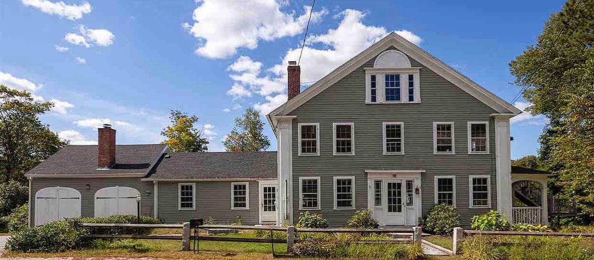 The exterior of a two-story house is presented, featuring a light green facade with white trim. Multiple windows adorn the front, allowing natural light to fill the interior. A spacious lawn with low greenery surrounds the entrance, enhancing the inviting appearance.