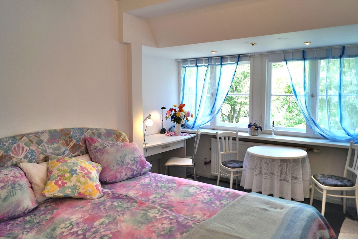 A bright room features a spacious bed adorned with colorful bedding and decorative pillows. A small table and chairs sit by a wide window draped with sheer, blue curtains, allowing natural light to fill the space. Fresh flowers add a touch of charm.