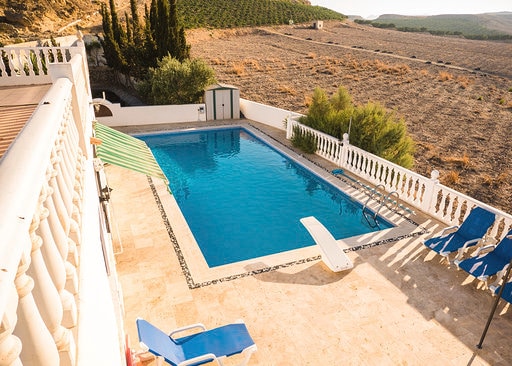 A large L-shaped swimming pool is positioned at the rear of the villa, surrounded by a spacious terrace with sun loungers. A diving board extends over the pool's edge, and scenic views of olive groves and mountains are visible in the background.