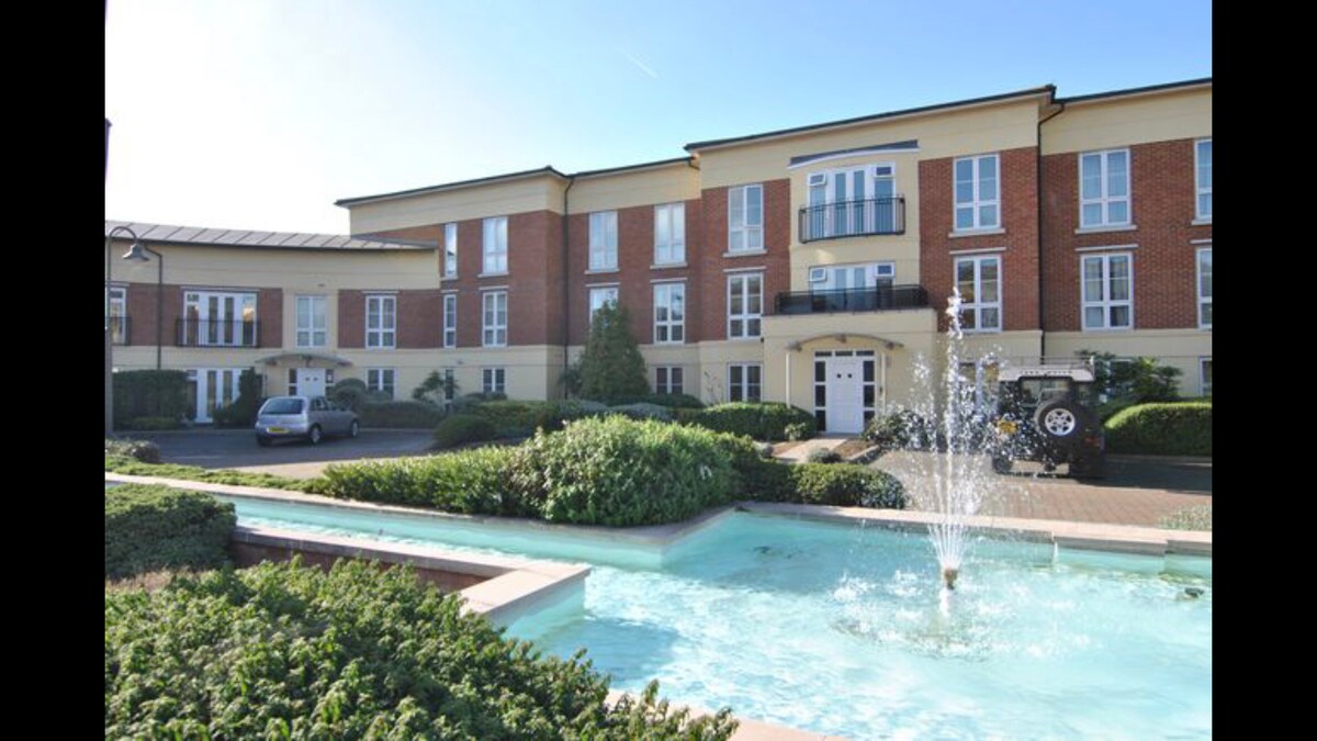 The building features a modern townhouse design with a light-colored façade, large windows, and a balcony. A tranquil fountain is seen in the foreground, surrounded by manicured greenery, creating a peaceful outdoor space. A parking area is located next to the building.