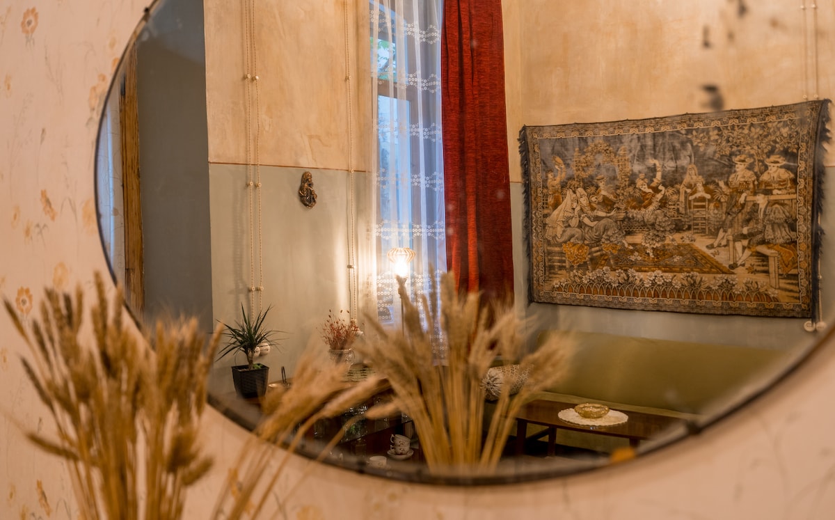 A round mirror reflects a cozy seating area adorned with a decorative tapestry. Soft light is provided by a nearby lamp, while dried plants are positioned in the foreground. The warm hues of the walls and curtains contribute to a welcoming environment.