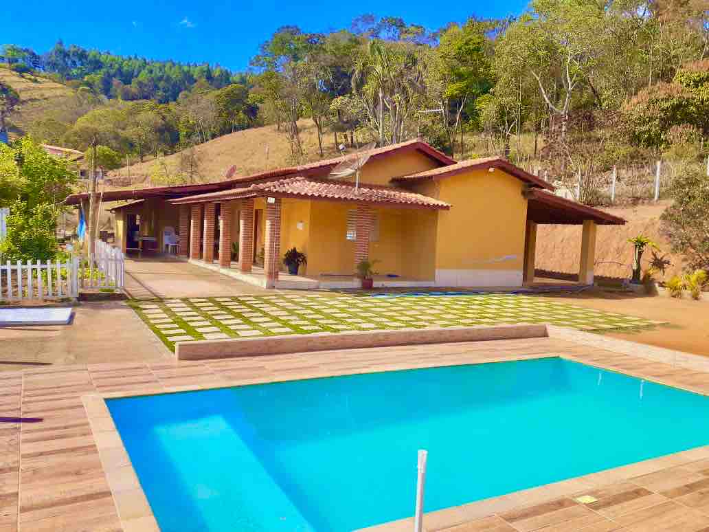 A spacious outdoor area features a clear swimming pool surrounded by tiled decking. A yellow building with a red-tiled roof and multiple windows is situated nearby, complemented by manicured green lawns and hills in the background.