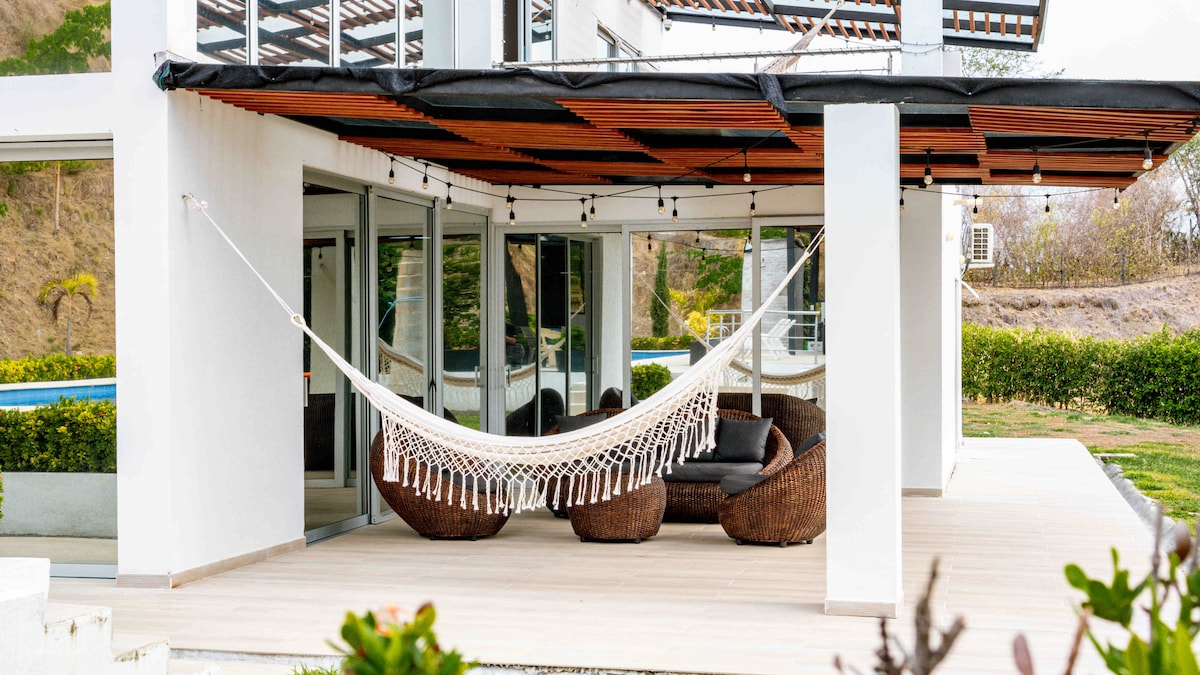 A spacious outdoor terrace is shown, featuring a relaxing hammock hung between two posts. Woven seating is present, offering a comfortable space to enjoy the view. Large glass doors provide visibility to the pool area and surrounding greenery, creating a serene environment.