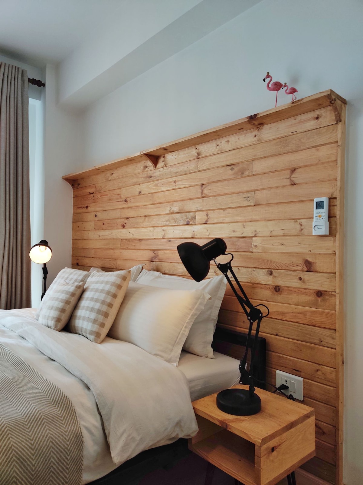 A cozy sleeping area is featured, showcasing a wooden accent wall. A spacious bed with white linens is complemented by two patterned pillows. Beside the bed, a wooden nightstand holds a lamp with a black lampshade, illuminating the space softly.