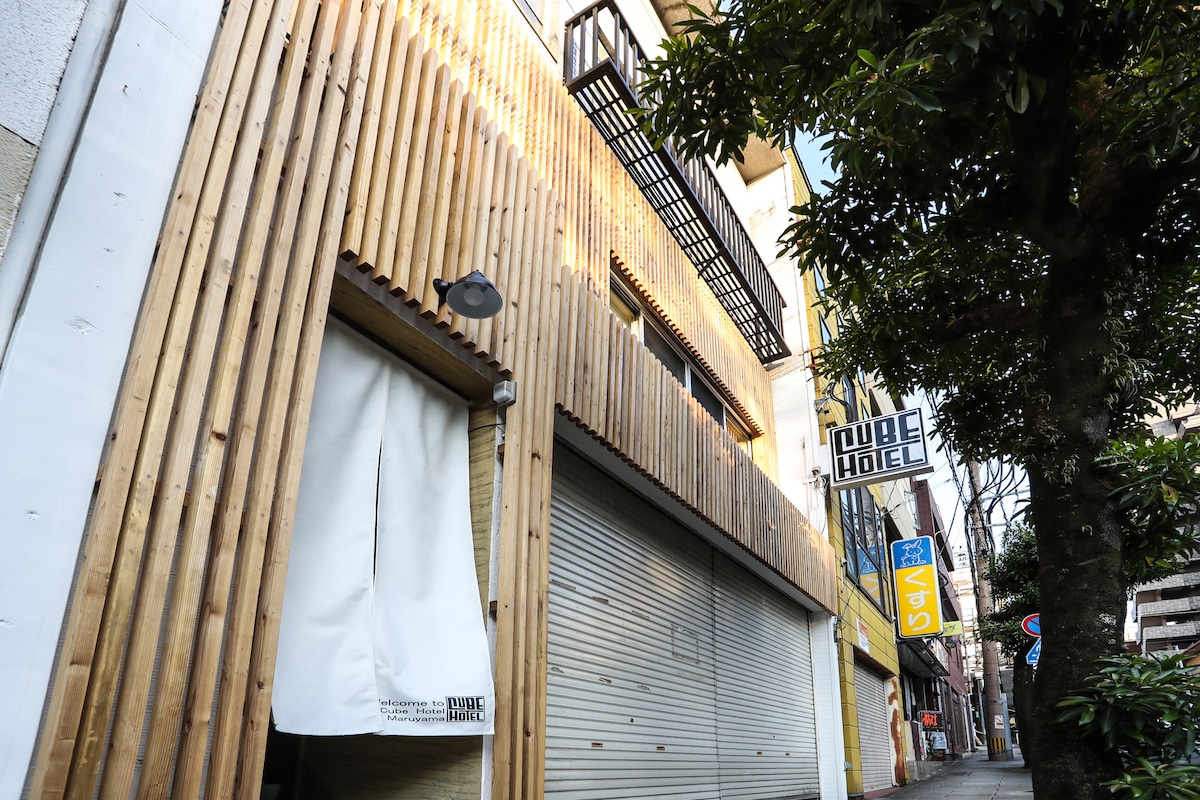 The exterior of the CUBEHOTEL MARUYAMA showcases a modern design with natural wooden cladding. Large windows allow for ample natural light, while a sign indicating the hotel's name is prominently displayed. A tree-lined street provides a welcoming atmosphere in the vicinity.